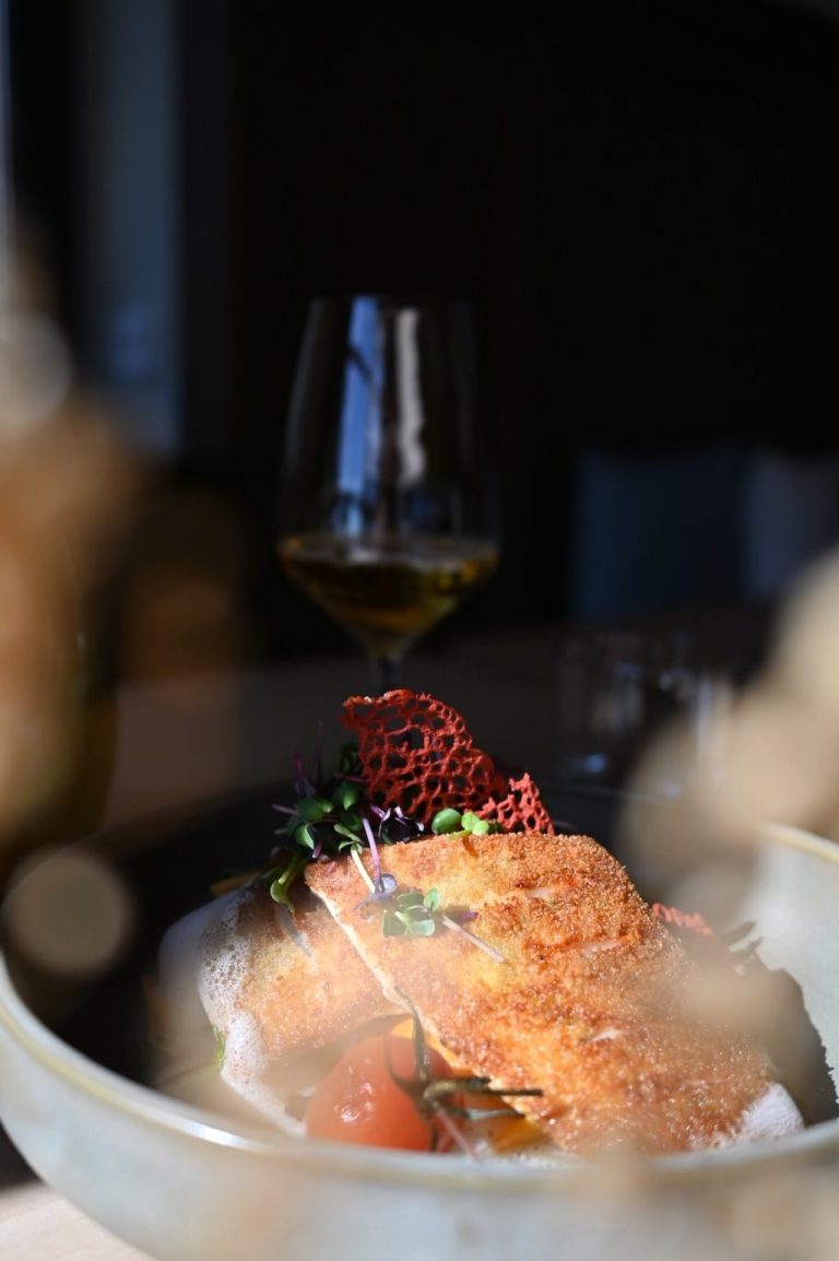 Gourmetgericht mit Fisch, Gemüse und einem Glas Wein im Hintergrund.